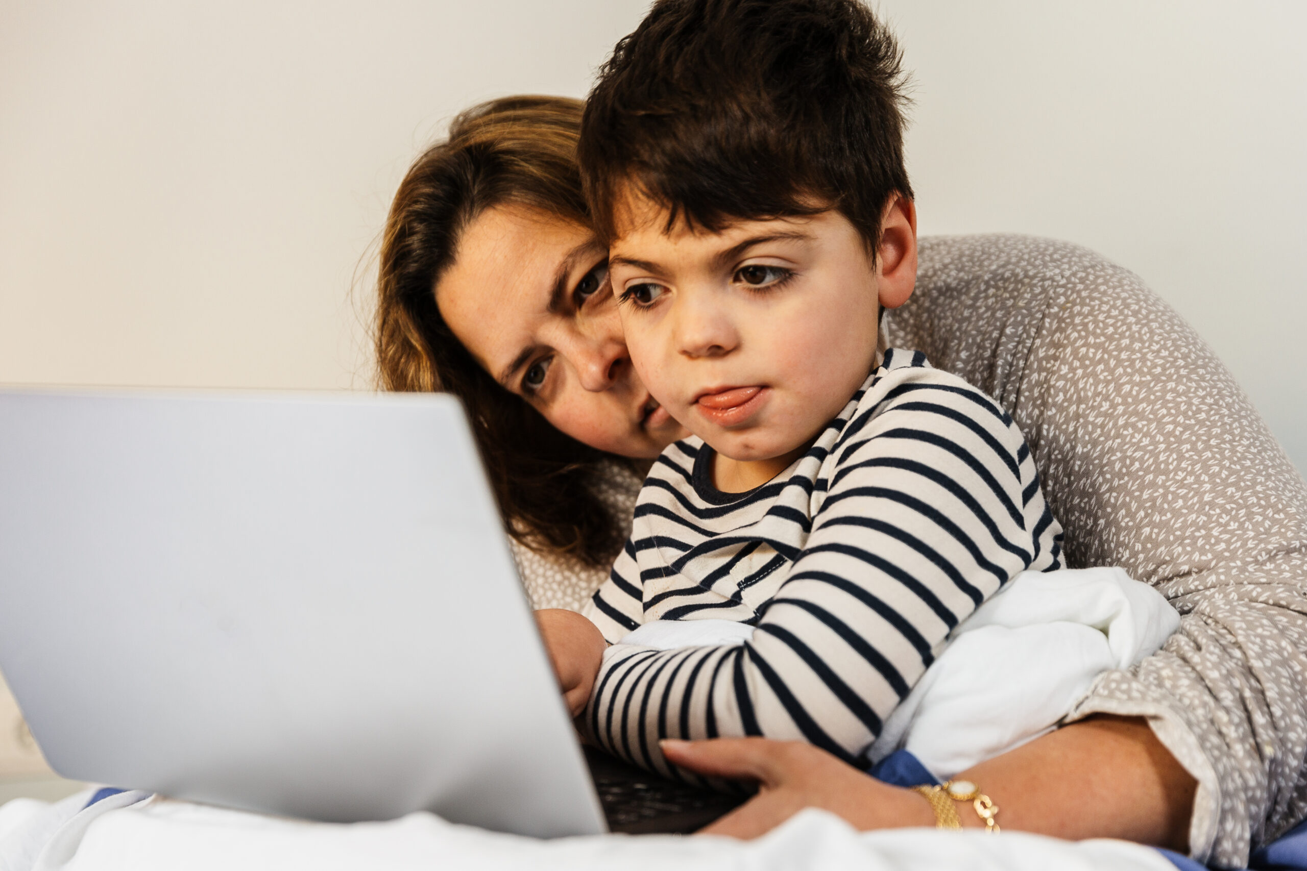 A five-year-old boy with a disability is with his mother in the parents' bedroom, sitting on the bed. They look at a laptop.